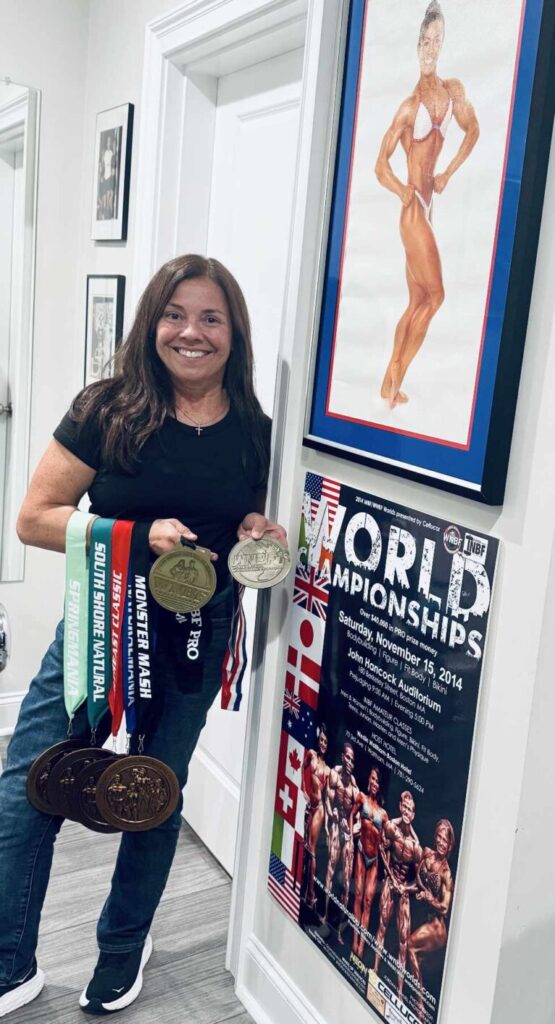 Nancy Andrews posing with medals from her natural bodybuilding events alongside a framed photo from her competitive career